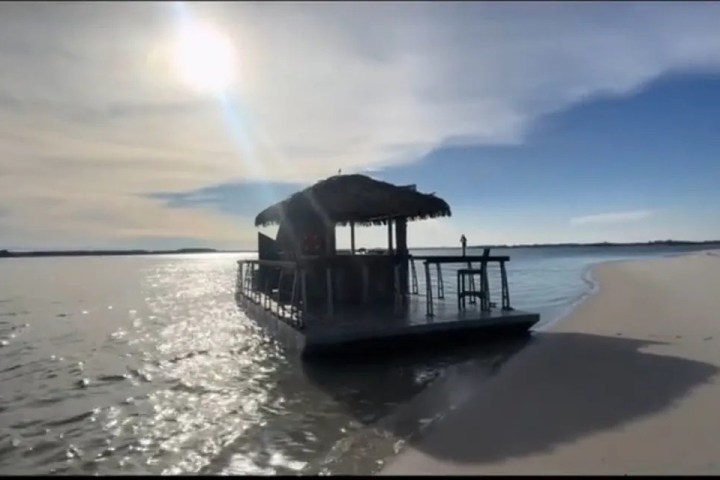 Floating tiki hut on calm water with sandy shore and bright sun.