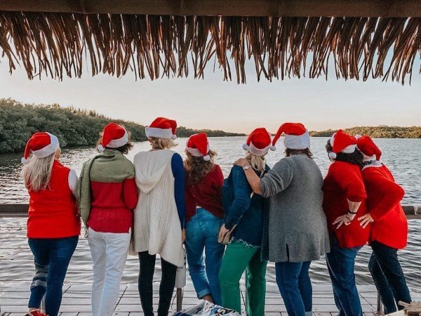 Seven people wearing Santa hats, back to camera, standing on a dock by the water.