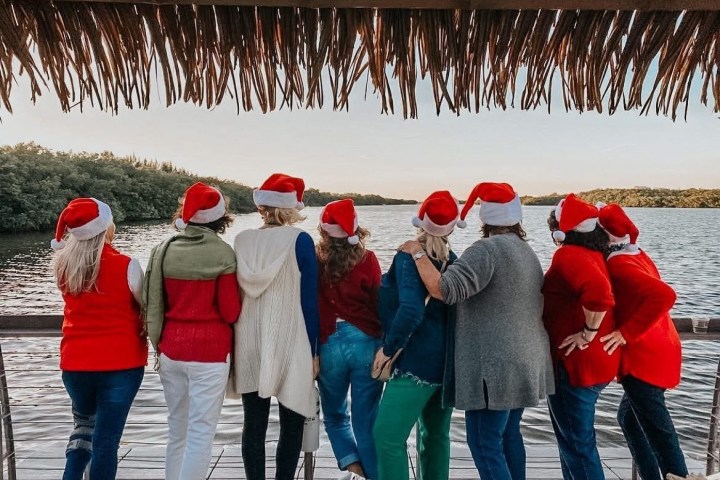 Seven people wearing Santa hats, back to camera, standing on a dock by the water.