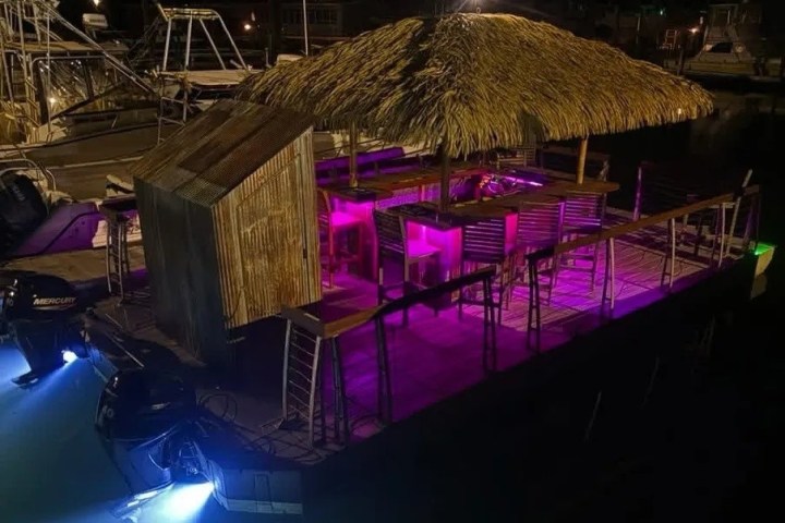 Night view of a thatched roof bar on a boat with purple lighting.