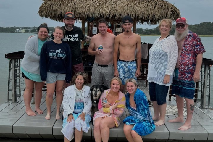 Group of people on a dock by the water, some in swimwear, with a thatched structure in the background.