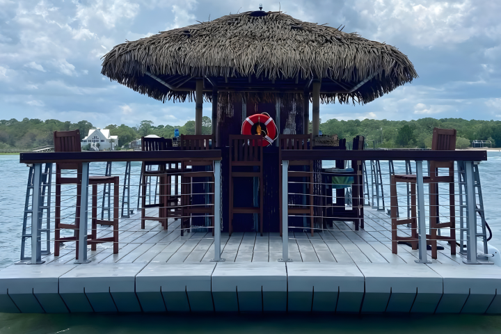 Floating tiki bar on water with thatched roof and wooden stools.