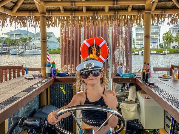 Child in captain's hat steering a boat under a tiki-style roof.