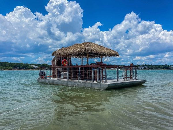 Floating tiki bar on calm water with cloudy sky.