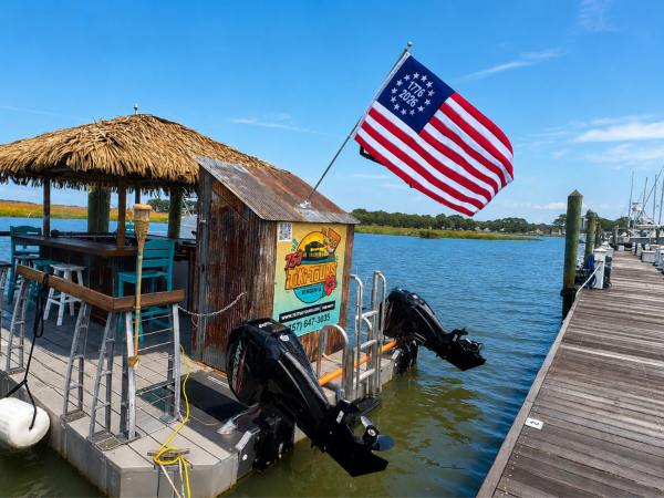 Floating tiki bar with bar stools and USA flag docked by a pier on a sunny day.