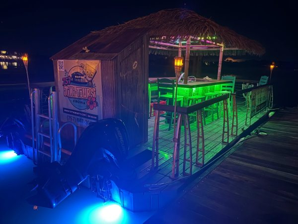 Illuminated tiki bar boat with colorful lights on water at night.