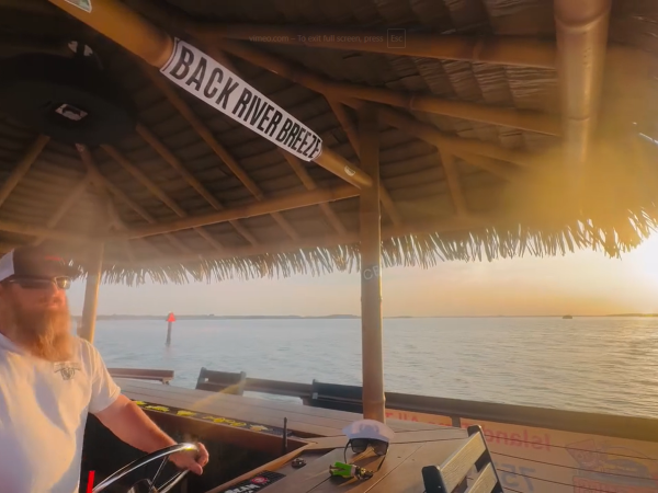 Bearded man steering boat under tiki hut roof at sunset.