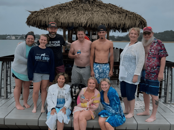 Group posing on a dock with a thatched roof hut near a body of water.
